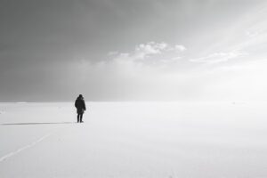 Een eenzame donkere figuur als ankerpunt in een uitgestrekt minimalistische zwart-wit sneeuwlandschap met dramatische lucht en lange schaduwen